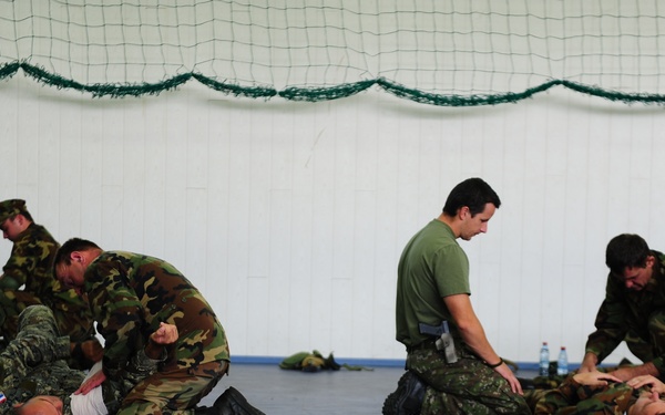 The image shows military personnel engaged in what appears to be combat training or hand-to-hand fighting drills inside an indoor facility. Several soldiers in camouflage uniforms are practicing ground fighting techniques, with some pinning others to the floor. An instructor in a green t-shirt observes and supervises the training session.