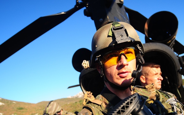 A group of military personnel in full combat gear are disembarking or assembling near a large helicopter, likely a CH-47 Chinook, in an open field with mountainous terrain in the background. The foreground soldier is prominently equipped with a tactical helmet, yellow-tinted protective eyewear, a communications headset, body armor, and is carrying a firearm. The scene appears to depict a military exercise or operational insertion.