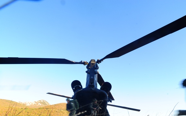 Military personnel in combat gear are rapidly boarding or disembarking a tandem-rotor helicopter, likely a CH-47 Chinook, on a grassy field. The image is captured from a low ground-level angle with motion blur on the soldiers, suggesting fast movement. The setting appears to be a mountainous terrain during early morning or late afternoon light.