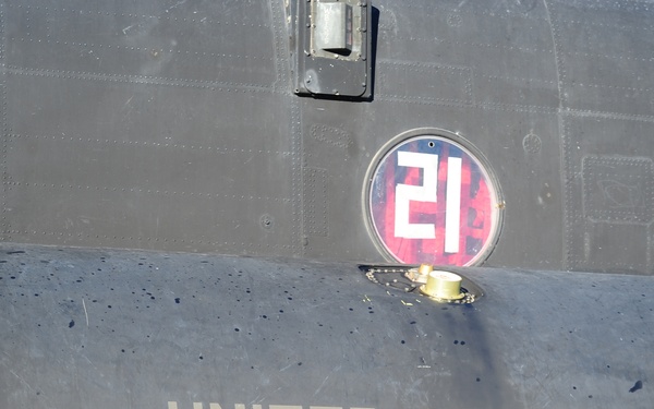 The image shows the fuselage of a United States Army military helicopter, likely a CH-47 Chinook or similar large transport helicopter, painted in olive drab/dark green. A circular insignia bearing the number '21' on a red and white background is visible on the aircraft's body. A shadow of a soldier carrying a weapon is cast on the lower left portion of the fuselage.