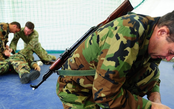 Military personnel in woodland camouflage uniforms are practicing combat casualty care or first aid techniques on a blue mat surface, likely in a training facility. The foreground shows a soldier with a rifle slung over his back applying a bandage wrap to another soldier's leg. A second pair of soldiers in the background are performing a similar medical aid drill.