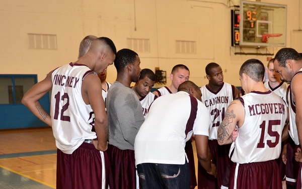 A basketball team huddles together on an indoor court, likely during a timeout or pre-game meeting. Players wearing white and maroon uniforms with visible names and numbers gather in a tight circle, with what appears to be a coach or team leader in street clothes addressing them. A scoreboard is visible in the background.
