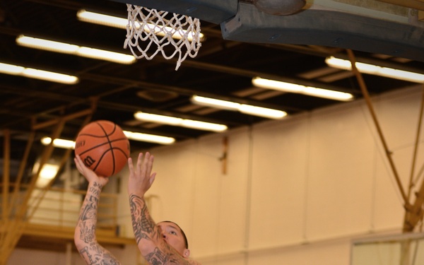 An indoor basketball game is in progress, with a player wearing a white 'Canyon #24' jersey attempting a layup or shot near the basket. Two defenders in blue 'Holloman' jerseys (numbers 33 and 22) are contesting the shot. The player shooting has extensive tattoos on both arms.