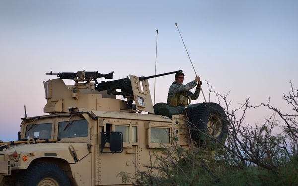 A military HMMWV (Humvee) is positioned in a desert scrubland environment at dusk or dawn. A soldier in military fatigues is seated in the gunner's turret position, appearing to adjust or handle radio antennas. The vehicle is equipped with a mounted weapon system and multiple communication antennas.