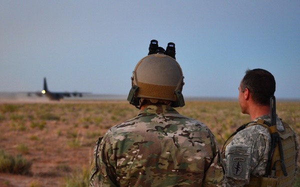 Two military personnel in different camouflage uniforms stand on red dirt terrain observing a large military transport aircraft (likely a C-130 Hercules) in the background. The soldier on the left wears MultiCam/OCP pattern with a tactical helmet equipped with night-vision goggle mounts, while the soldier on the right wears Army Combat Uniform (ACU) digital pattern with visible Special Forces insignia. The setting appears to be an austere airstrip in an arid environment, likely in Africa based on the red soil and sparse vegetation.