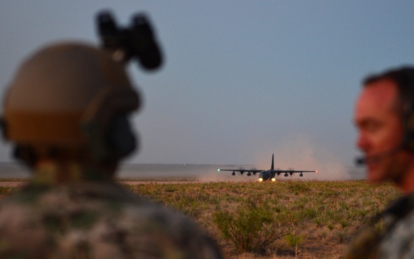 Two military personnel in camouflage uniforms observe a large military transport aircraft, likely a C-130 Hercules, landing or taking off on an unpaved airstrip in an arid, scrubland environment at dusk or dawn. The foreground figures are out of focus while the aircraft in the background is the focal point, kicking up dust during its ground roll. The scene appears to depict a tactical airfield operation or training exercise.
