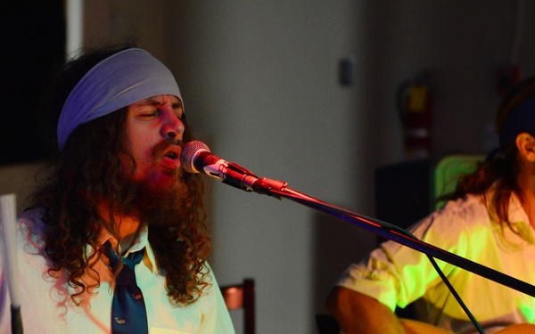 Two male musicians are performing live on stage under colorful lighting. The foreground performer has long curly hair and a beard, wearing a blue headband and teal tie, singing into a microphone while playing conga drums with drumsticks. The background performer, similarly dressed with a blue headband, plays an acoustic guitar and sings into a separate microphone.