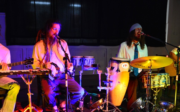 A four-piece band performs live in what appears to be an indoor venue with colorful stage lighting. The musicians are playing a mix of string and percussion instruments, all seated or positioned close together on a wooden floor stage. The group has a casual, bohemian aesthetic with long hair and relaxed attire.