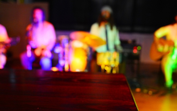 A live music performance is taking place in a dimly lit venue with colorful stage lighting. In the foreground, two red promotional flyers for a band called 'Full Service' advertising a 'Free Show' are placed on a dark wooden table. The background shows approximately four musicians performing on stage, including a guitarist, drummer, and other instrumentalists.