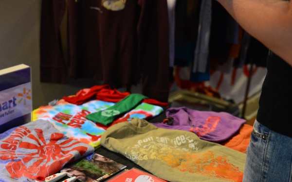 A merchandise or promotional display table covered with a black tablecloth, featuring various folded t-shirts in multiple colors and promotional materials. A Walmart-branded sign is prominently displayed on the left side of the table. A person wearing jeans is standing to the right, browsing the items.