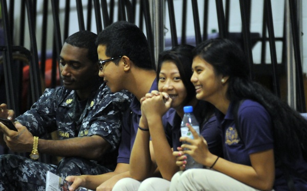 A group of four people are seated on bleachers in what appears to be a gymnasium or indoor sports venue. A military servicemember in Navy digital camouflage uniform is showing something on a smartphone to three young people wearing matching navy blue polo shirts and khaki pants, likely students in a school uniform. The group appears engaged and cheerful, with two of the students smiling.