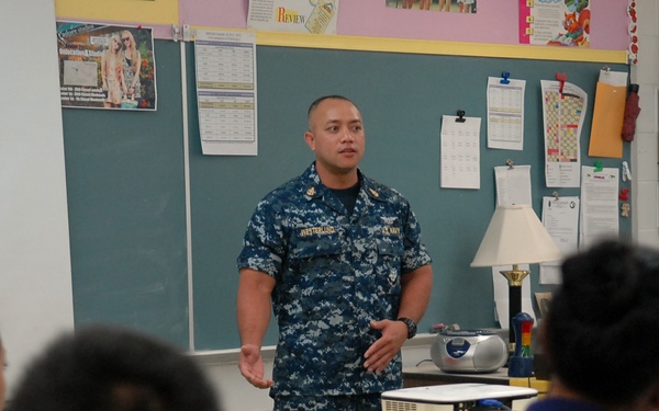 A U.S. Navy sailor in blue digital camouflage (NWU Type I) uniform is addressing a classroom of students who are seated at desks. The setting appears to be an elementary or middle school classroom, with bulletin boards, a chalkboard, and educational materials visible in the background. This appears to be a community outreach or career education visit by a Navy service member.