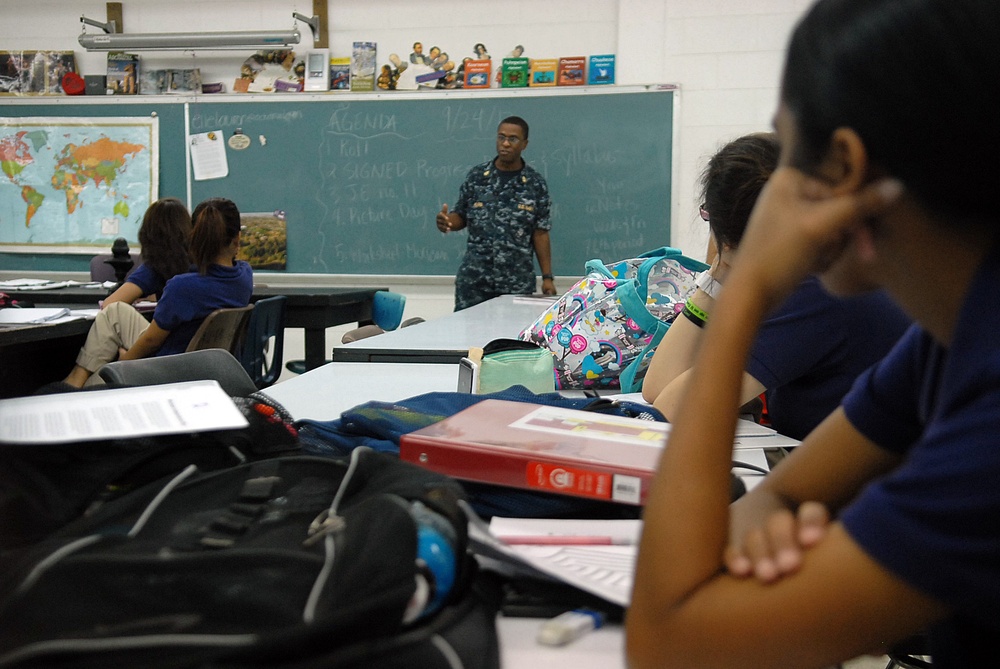 A person in U.S. Navy digital camouflage uniform stands at the front of a classroom addressing students seated at desks. A chalkboard behind them displays an agenda dated '1/24' with several numbered items listed. The classroom setting includes typical school elements such as maps, books, and student backpacks.