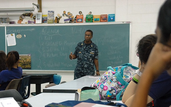 A person in U.S. Navy digital camouflage uniform stands at the front of a classroom addressing students seated at desks. A chalkboard behind them displays an agenda dated '1/24' with several numbered items listed. The classroom setting includes typical school elements such as maps, books, and student backpacks.