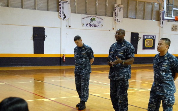 Four U.S. Navy personnel in blue digital camouflage (NWU Type I) uniforms are standing on a gymnasium floor, apparently addressing a seated audience of students. The setting appears to be a school gymnasium, likely George Washington High School based on the 'Geckos' and 'George Washington Cheer' banners visible on the walls. One service member appears to be speaking into a microphone, suggesting a recruitment or community outreach presentation.