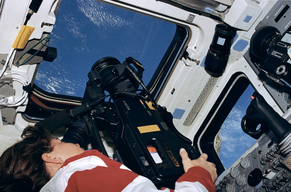STS-93 MS Coleman aims a HD video camera through a window on the flight deck