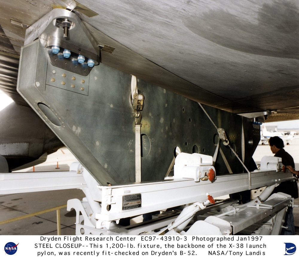 A NASA photograph from January 1997 taken at Dryden Flight Research Center showing a close-up of a 1,200-lb steel fixture that serves as the backbone of the X-38 launch pylon, fit-checked on a B-52 aircraft. The image shows the underside of the B-52 with the large metallic pylon structure attached, along with support equipment and a technician. The photo is credited to NASA photographer Tony Landis.