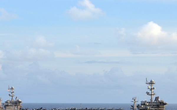 The image shows two large US Navy aircraft carriers operating in open ocean waters. The foreground carrier displays the hull number '73' on its island superstructure, identifiable as USS George Washington (CVN-73). Both flight decks are loaded with aircraft, and a helicopter is visible in the background sky to the left.