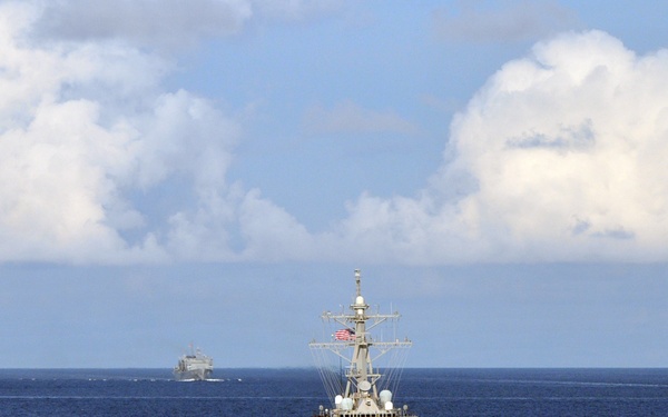 The image shows a U.S. Navy carrier strike group underway at sea, consisting of multiple warships in formation. A guided-missile destroyer is prominently positioned in the foreground, flying an American flag, flanked by two aircraft carriers on the left and right background. A smaller vessel, possibly a supply or amphibious ship, is visible in the center background.