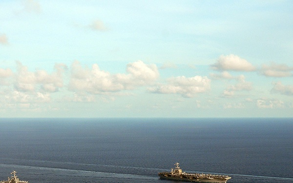 An aerial photograph showing a naval carrier strike group formation underway at sea. The formation includes at least two aircraft carriers flanked by multiple escort vessels including destroyers or cruisers. The ships are sailing in a coordinated formation across open ocean under partly cloudy skies.