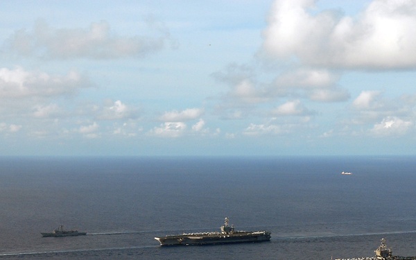 An aerial photograph shows a naval carrier strike group formation underway at sea. The formation includes two large aircraft carriers flanked by smaller escort vessels including cruisers and destroyers. The ships are leaving visible white wakes as they transit across calm, deep blue ocean waters.