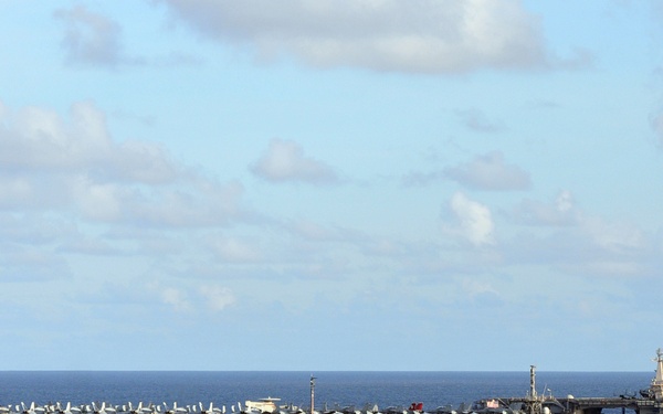 The image shows a naval formation at sea featuring two large US Navy aircraft carriers and a smaller escort warship sailing in open ocean. The carrier on the left bears the hull number '73' and the one on the right bears '74', both with aircraft visible on their flight decks. A smaller frigate or destroyer-class vessel with a partial hull number ending in '8' is positioned between and in front of the carriers.