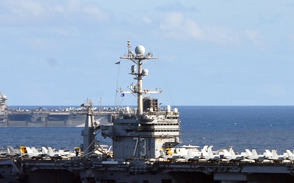 The image shows two U.S. Navy aircraft carriers operating together at sea in open blue water. The foreground carrier bears the hull number '73', identifiable as USS George Washington (CVN-73), a Nimitz-class nuclear-powered aircraft carrier. A second carrier is visible in the background to the port side, with aircraft visible on both flight decks.