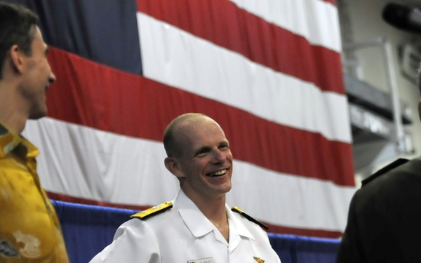 A U.S. Navy officer in white dress uniform is smiling and conversing with a civilian wearing a colorful patterned shirt, in front of a large American flag backdrop. The setting appears to be an official military ceremony or event held indoors. A third person, partially visible on the right, appears to be operating a camera or recording equipment.
