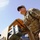 A Kentucky soldier supervises weapons training in southern Afghanistan A U.S. military soldier wearing MultiCam (OCP) camouflage uniform and tactical vest is handling green military ammunition cans on or near a tan-colored military vehicle. The photo is taken from a low angle looking upward, with a clear blue sky in the background. Patches on the uniform indicate ISAF/U.S. Army affiliation and a 'SAPPER' tab is visible.