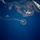 An orbital or high-altitude aerial photograph showing a volcanic island actively emitting a plume of smoke or ash, drifting westward across the ocean. A larger landmass with rugged, mountainous terrain is visible in the upper portion of the image, along with several smaller islands. The deep blue ocean contrasts sharply with the brown-green landmasses and the white volcanic plume.