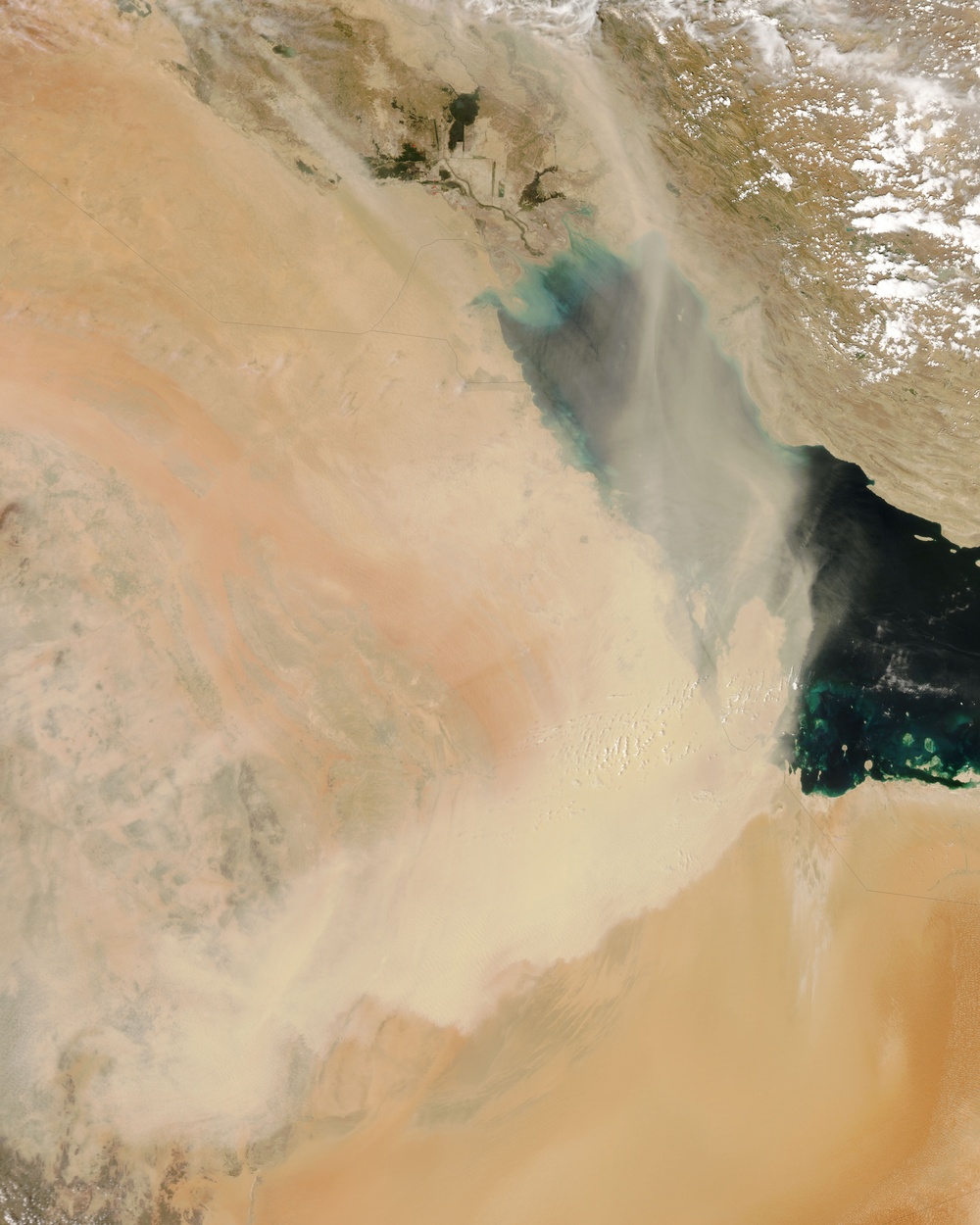 This is a satellite image showing a large dust or sand storm sweeping across an arid desert region toward a body of water, likely the Persian Gulf area based on the geography. A massive plume of pale dust obscures much of the land surface, with darker smoke or aerosol layers visible above it. The image captures a significant atmospheric event over what appears to be the Arabian Peninsula and surrounding waters.