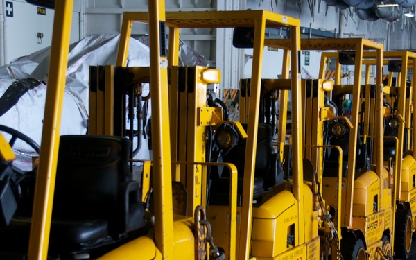 A long row of yellow Hyster 60 forklifts are parked in a line inside what appears to be a large industrial facility or ship's cargo hold. The forklifts are secured with chains and appear to be stored or staged for deployment. The uniform yellow color and identical model suggest these are fleet vehicles belonging to a single organization.