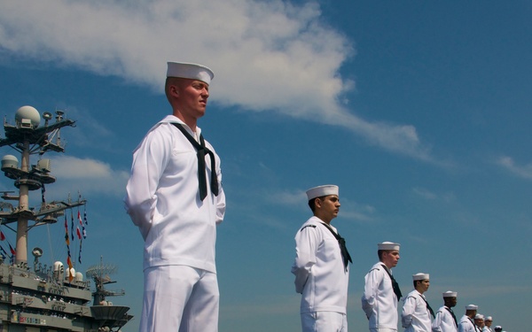 A long line of U.S. Navy sailors in dress white uniforms are standing at attention on the flight deck of an aircraft carrier, performing a 'manning the rails' ceremonial formation. The sailors are evenly spaced along the edge of the flight deck, with the ship's island superstructure visible in the background to the left. The scene appears to be a formal naval ceremony, likely a port arrival or departure.