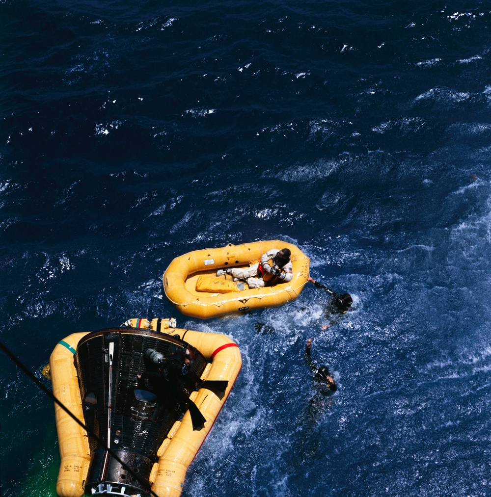An aerial photograph shows a space capsule recovery operation at sea. A person in a white spacesuit is seated in a yellow inflatable life raft, while swimmers in the water assist nearby. A dark, cone-shaped space capsule with yellow flotation collar is visible in the lower left corner of the frame.
