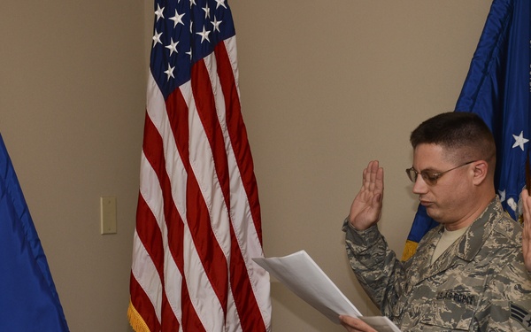 Three U.S. Air Force personnel in Airman Battle Uniform (ABU) camouflage are participating in a formal military oath ceremony indoors. The central figure is reading from a document while the other two individuals have their right hands raised, indicating they are taking an oath. The ceremony is conducted in front of an American flag and Air Force flags.