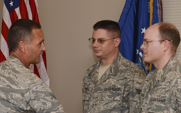 Three U.S. Air Force personnel in Airman Battle Uniform (ABU) camouflage are gathered in what appears to be an official military ceremony. Two of the airmen are shaking hands while a third observes, suggesting a formal recognition or promotion event. An American flag and a blue Air Force unit flag are visible in the background.