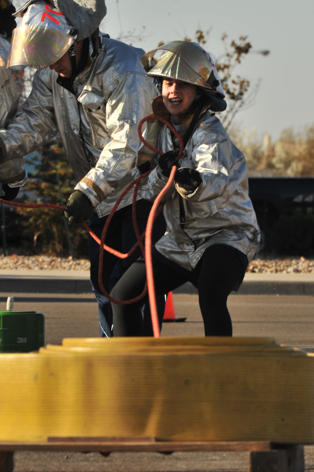 Two individuals wearing silver aluminized proximity/fire-resistant suits and helmets with face shields are engaged in what appears to be a firefighting training exercise. They are handling a red hose while crouching near yellow cylindrical equipment, likely a fire hydrant or training prop. The setting appears to be an outdoor training area with a paved surface.