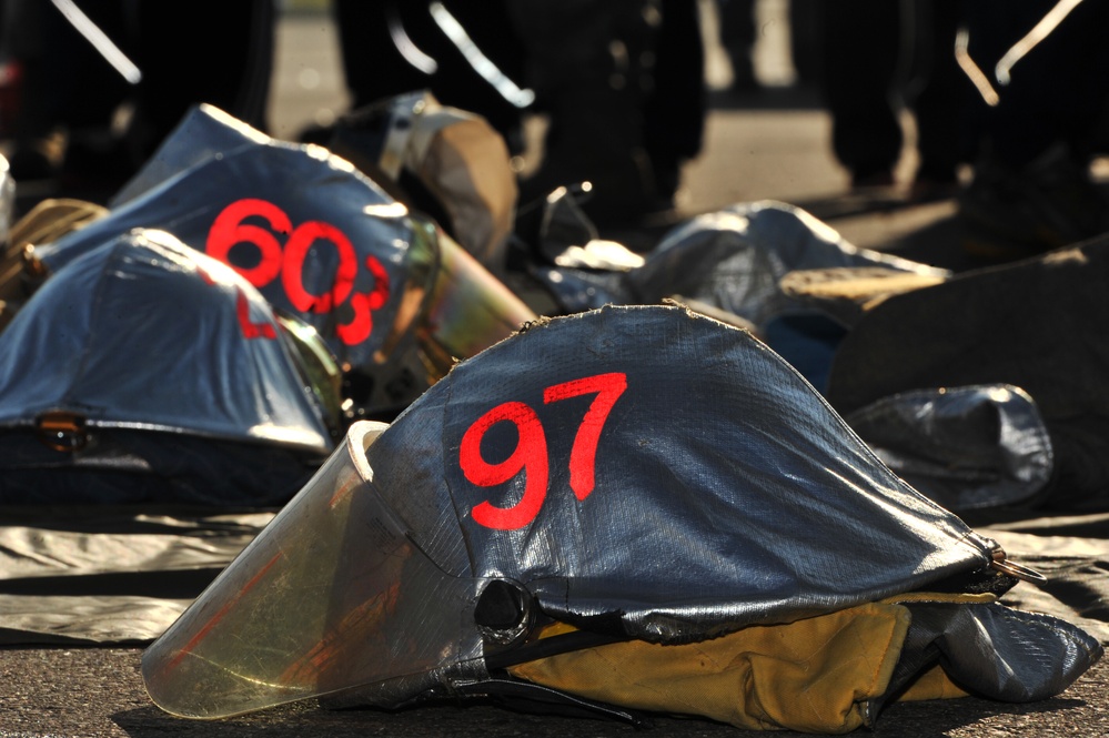 The image shows multiple firefighter or emergency responder helmets laid out on a ground surface, each marked with red numbers. The foreground helmet is numbered '97' and a background helmet shows '603'. Personnel in dark uniforms are visible standing in the background.