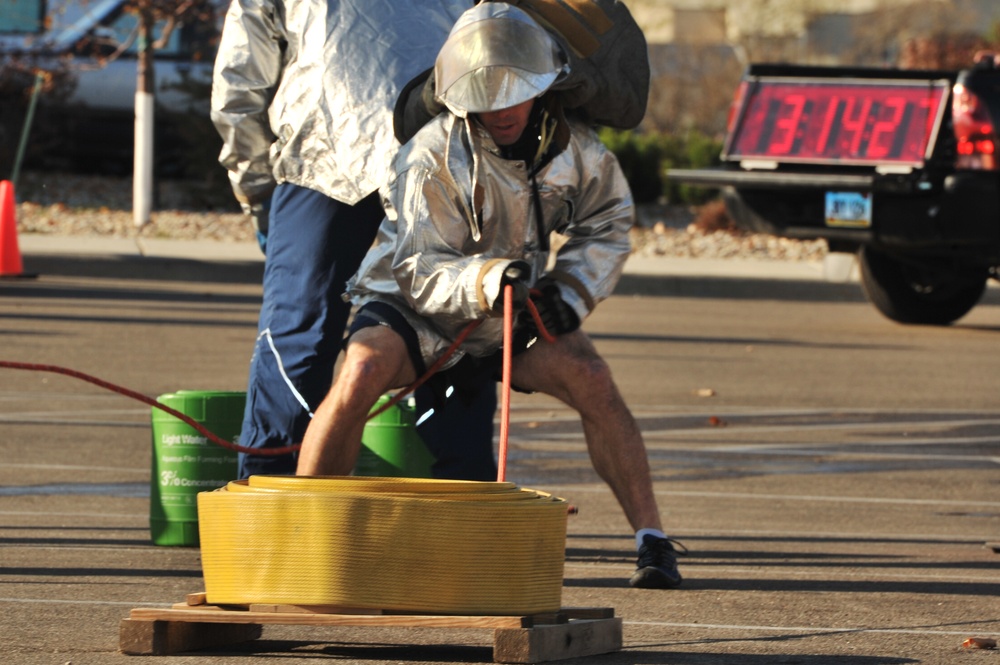 Two individuals wearing silver/aluminized proximity suits and helmets are participating in what appears to be a firefighter competition or training exercise in a parking lot. The foreground person is actively pulling a rope or hose connected to a coiled yellow fire hose on a wooden pallet. A digital timer in the background displays '13:14:21', suggesting this is a timed competitive event.