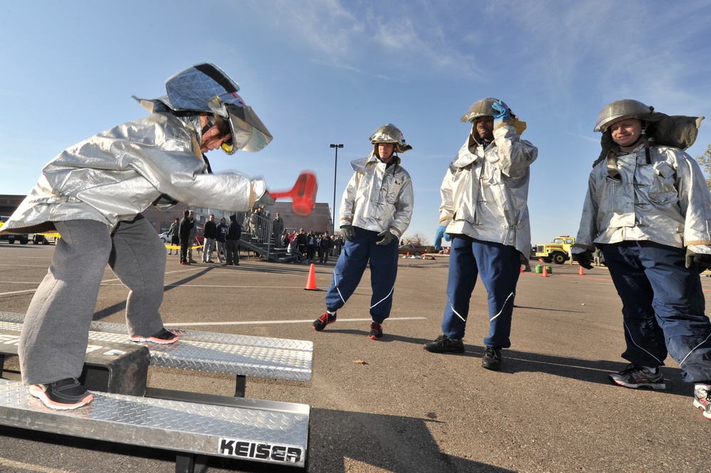 A group of individuals in silver/aluminized proximity firefighting gear are participating in what appears to be a firefighting training exercise or competition in a parking lot. One person is actively using a red sledgehammer on a Keiser Force Machine, a standard firefighting fitness/skills testing apparatus. Three other participants in similar gear stand nearby observing, with a crowd and emergency vehicles visible in the background.
