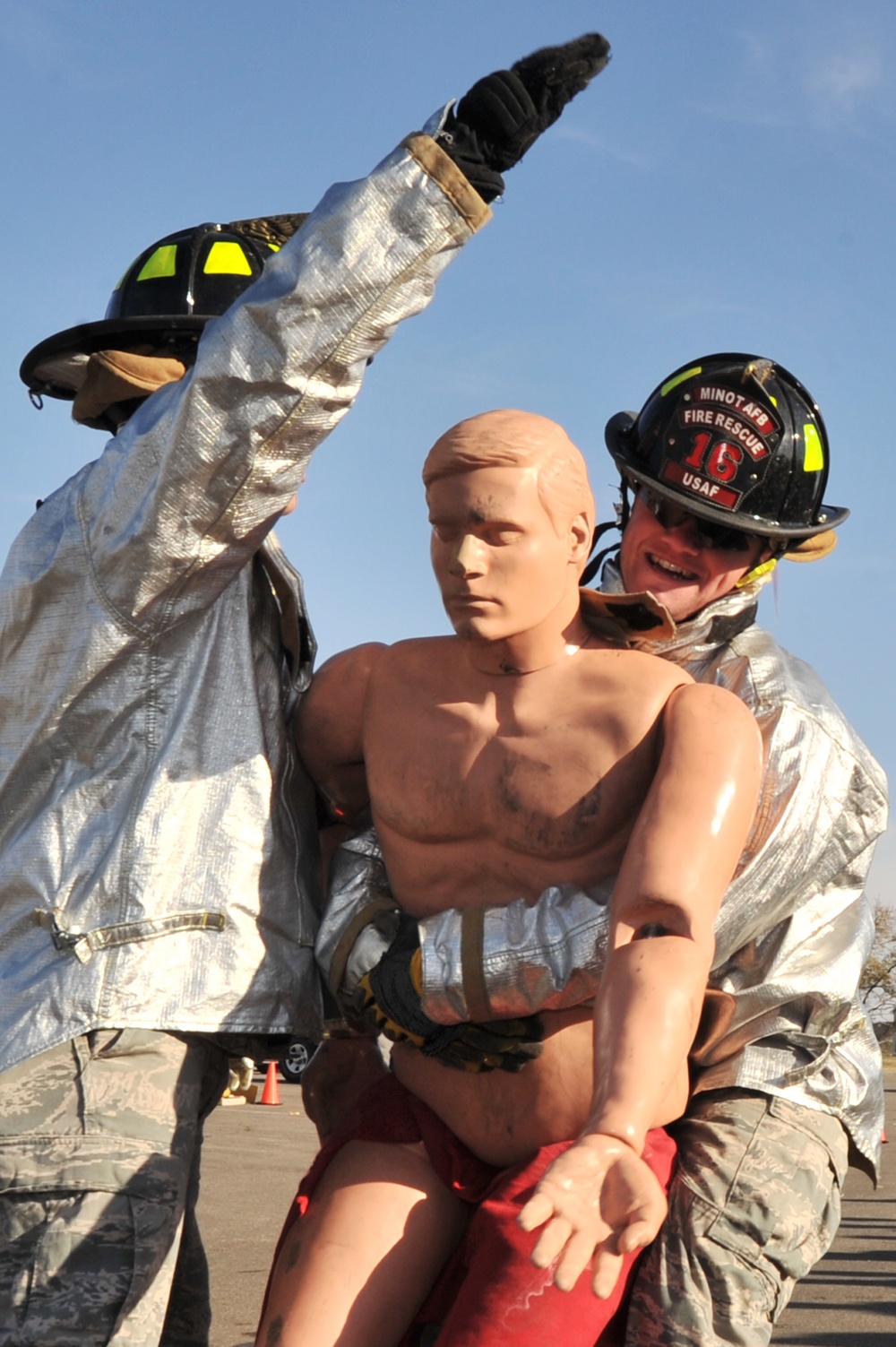 Two firefighters in silver/aluminized proximity suits and helmets are conducting a rescue training exercise, carrying a full-size human rescue mannequin. The firefighter on the right wears a helmet marked 'Minot AFB Fire Rescue 16 USAF', identifying this as a United States Air Force fire rescue training drill. The exercise appears to be taking place on a paved outdoor surface under clear skies.