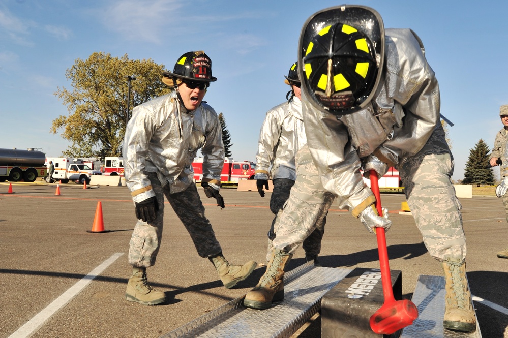 Military firefighters in proximity/aluminized protective suits are performing a training exercise on a paved surface, likely at an Air Force base. One firefighter is striking a metal object with a red sledgehammer while another in a black firefighter helmet cheers or shouts encouragement. Fire trucks and orange traffic cones are visible in the background.