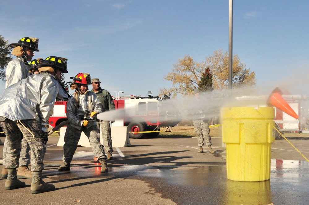 A group of military personnel in camouflage uniforms and firefighting gear are conducting a fire hose training exercise on a paved surface. Several individuals wearing silver proximity suits and firefighting helmets are operating a pressurized hose aimed at a large yellow container with an orange cone on top. A red fire truck is visible in the background, suggesting this is an organized training drill at a military installation.