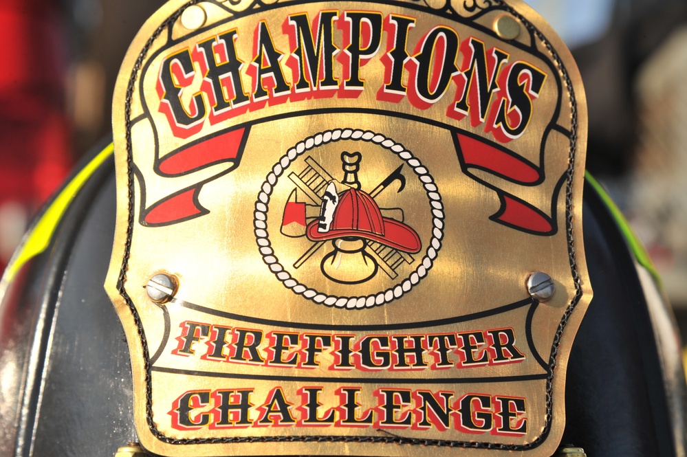 The image shows a close-up of a gold championship belt or award plaque for a 'Firefighter Challenge' competition. The ornate gold plate features the word 'CHAMPIONS' at the top with red ribbon banners, a central circular emblem depicting firefighting equipment, and 'FIREFIGHTER CHALLENGE' text at the bottom. The plaque appears to be mounted on a dark leather or vinyl backing secured with visible screws.