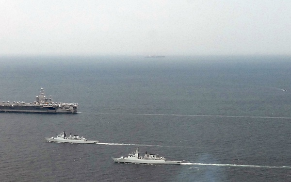 An aerial photograph shows four naval vessels sailing in a loose formation across open ocean. The formation includes one large aircraft carrier in the background and three smaller warships — likely destroyers or frigates — in the foreground. At least one vessel is emitting visible exhaust smoke, suggesting active propulsion at speed.