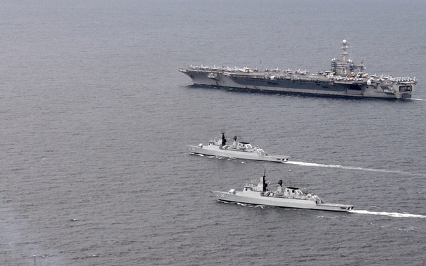 An aerial photograph shows four naval vessels sailing in a loose formation on open ocean. The formation includes one large aircraft carrier in the background and three smaller warships — two frigates or destroyers in the middle and one frigate in the foreground. The ships appear to be conducting a joint naval exercise or transit operation.