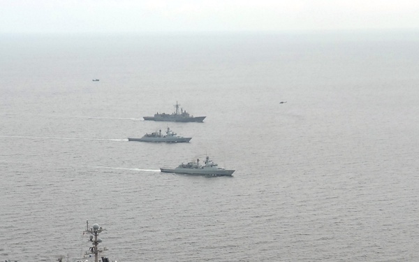 An aerial photograph shows a large aircraft carrier bearing hull number '73' in the foreground, accompanied by three smaller escort vessels in formation behind it. The ships are underway on open, calm ocean waters under an overcast sky. The carrier's flight deck is visibly populated with numerous aircraft arranged in rows.