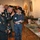 The image shows a military or veterans' exhibit/event where two U.S. Army soldiers in dress uniforms are conversing with an elderly civilian man wearing a black cowboy hat and blue jeans. A display table in the foreground contains WWII-era military memorabilia including a U.S. Marines recruitment poster, a military helmet on a mannequin head, canteen, and other equipment. An American flag and framed artwork are visible on the walls in the background.