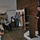 A U.S. Army officer in dress uniform is delivering a speech at a wooden podium during what appears to be a military or veterans' ceremony. An American flag is displayed prominently behind him, and an audience of mixed ages is seated and standing to his left. The event appears to be held in a rustic indoor venue with wood-paneled walls.