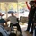 Two U.S. Army soldiers in dress uniforms are standing and conversing with elderly civilians, likely veterans, at what appears to be a Veterans Day or military appreciation event at a care facility or community center. The setting includes a covered porch or sunroom with large windows overlooking a parking area with autumn trees. The interaction appears respectful and ceremonial in nature.